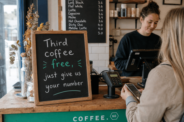 A handwritten chalkboard sign next to a coffee till reading 'Third coffee free, just give us your number'